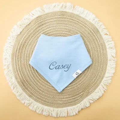 Light blue bib with 'Casey' embroidery on a round woven mat with tassels.