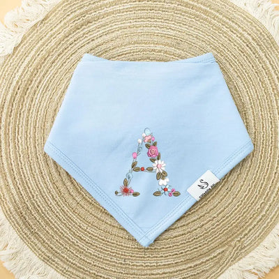 Light blue baby bib with floral letter 'A' embroidery on a woven mat