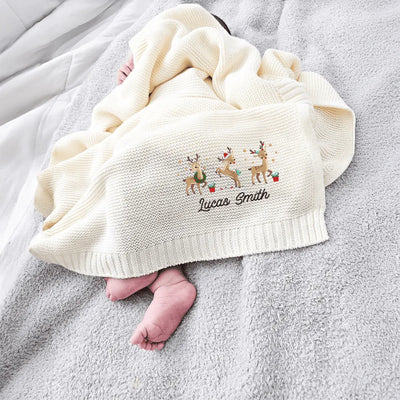 Baby wrapped in a cream personalized blanket with cute embroidered reindeer and text, lying on a gray surface.