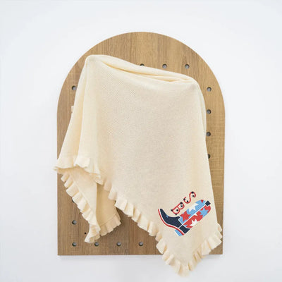 Cream personalized baby blanket with a colorful usa star cowboy boots logo on a wooden board against a white background
