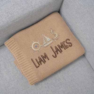 Personalized baby blanket with anchor and sailboat embroidered design and 'LIAM JAMES' text on a gray surface