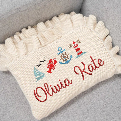 Cream personalized baby blanket with nautical-themed embroidery and 'Olivia Kate' text on a gray surface.